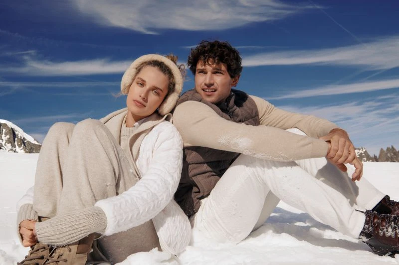 Couple seated in snow wearing winter clothing, including earmuffs, sweater, vest, and boots, mountains and blue sky in background.