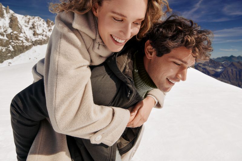 Couple smiles in snowy mountain landscape. Woman rides piggyback on man in winter coats with bright sky and mountain range backdrop.