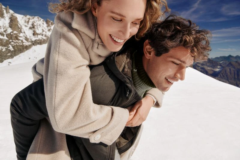 Couple smiles in snowy mountain landscape. Woman rides piggyback on man in winter coats with bright sky and mountain range backdrop.