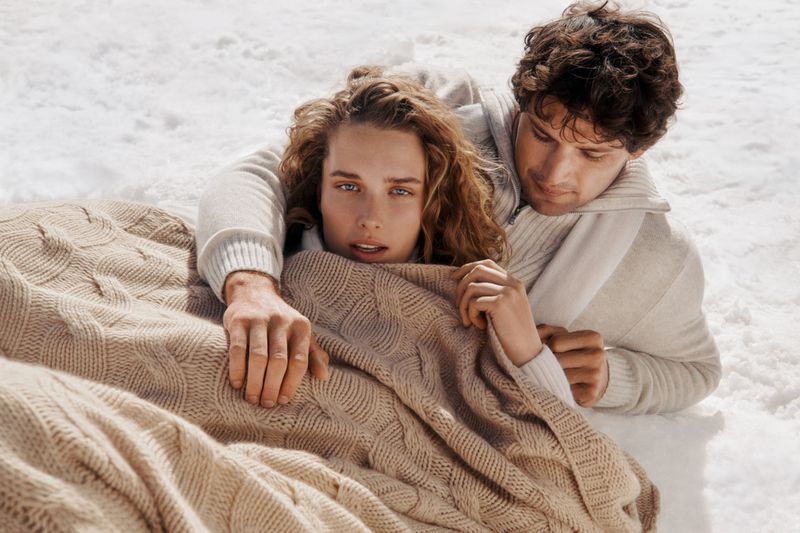 A serene scene of a couple lying in the snow under a knitted blanket, both wearing neutral-toned sweaters, capturing a cozy, intimate moment.