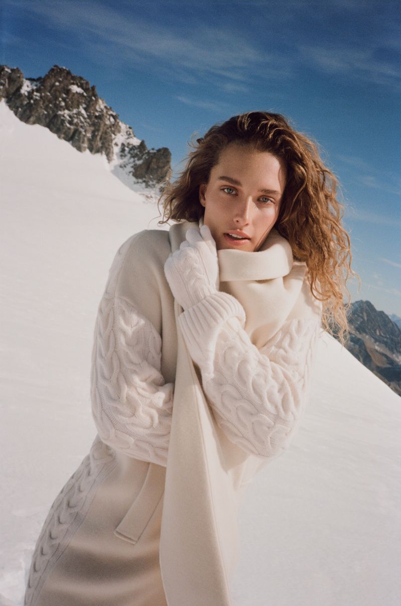 A woman with wavy hair wears a cream cable-knit sweater and scarf in a snowy mountain landscape under a bright blue sky.