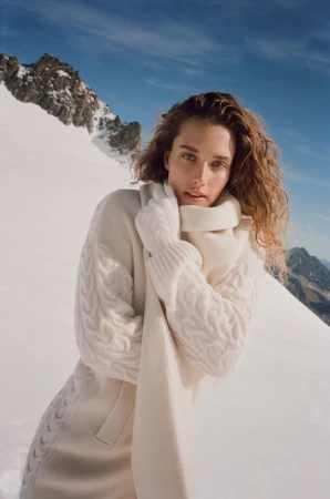 A woman with wavy hair wears a cream cable-knit sweater and scarf in a snowy mountain landscape under a bright blue sky.