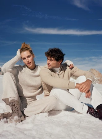 A man and a woman in neutral-colored winter wear sit on a snow-covered mountain, under a clear blue sky.