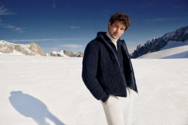 Man wearing a navy zip jacket and white trousers standing on a snowy mountain landscape under a clear blue sky