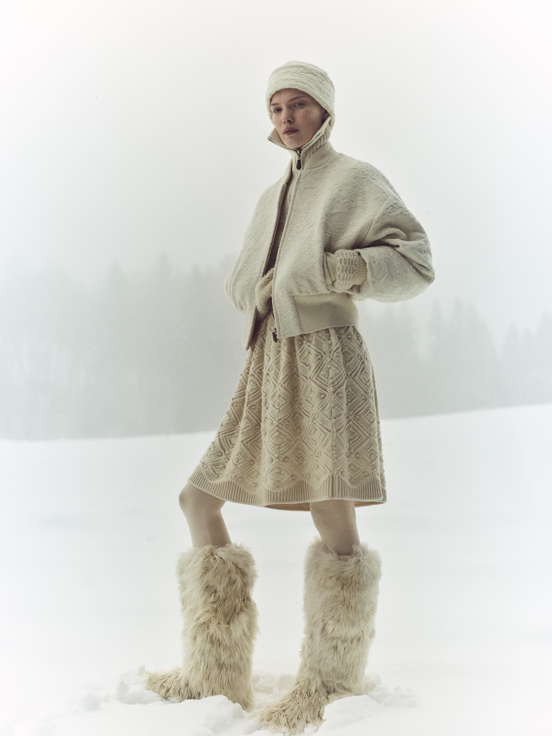 Adult female fashion model poses in an outdoor winter setting, wearing a light-colored knit skirt, jacket, hat and striking furry boots.