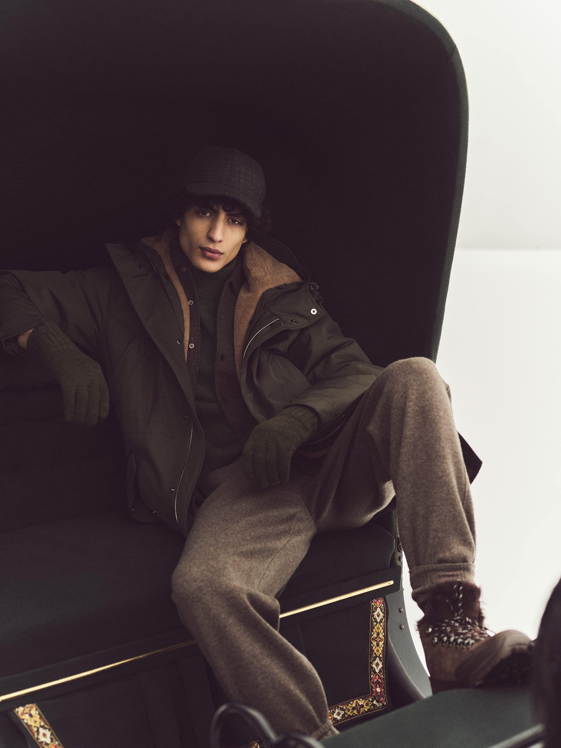 A stylish young man in a dark cap and olive jacket lounges in a carriage-like seat, showcasing a fashionable, relaxed pose and muted tones.