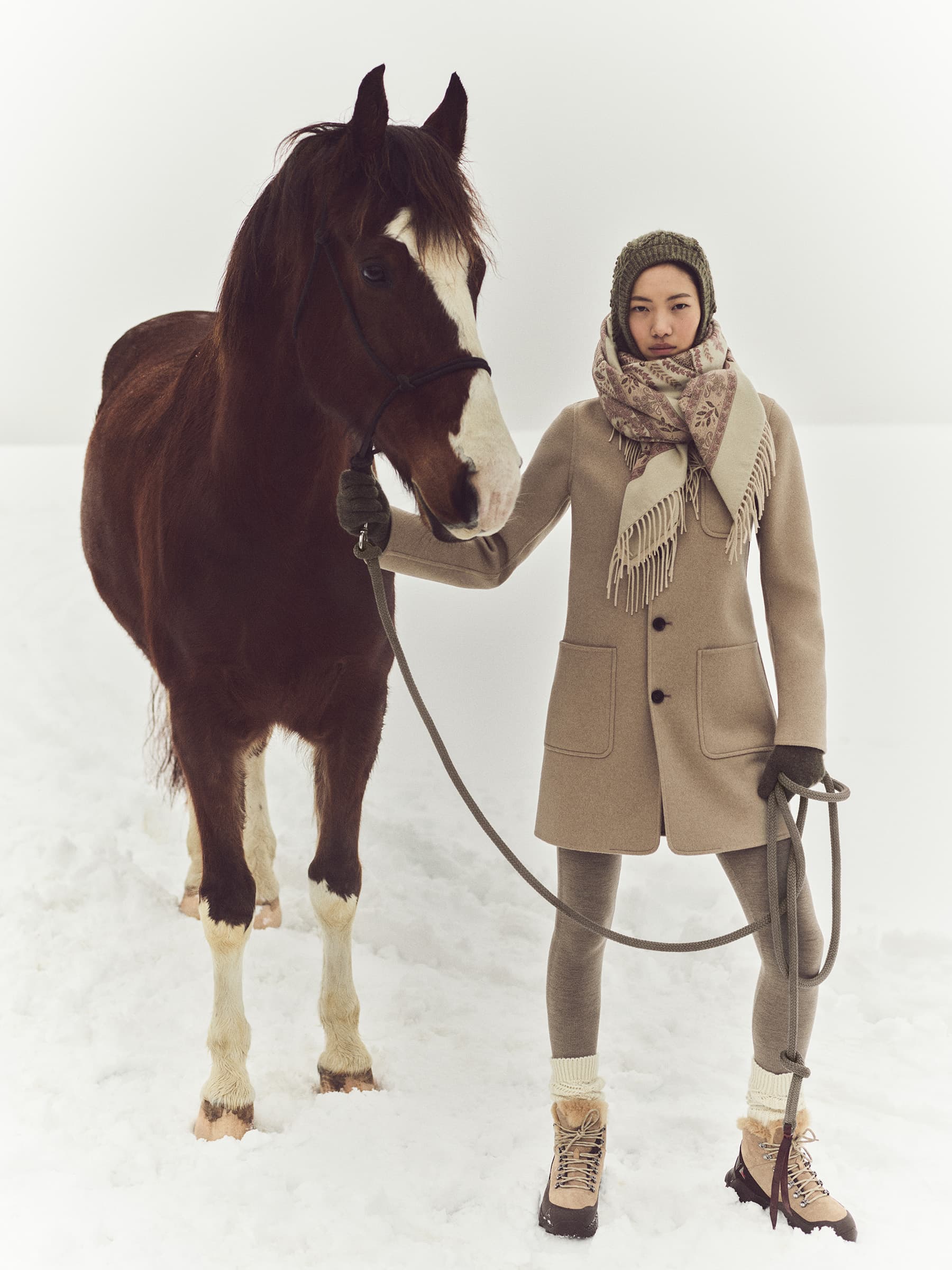 Woman in coat poses with brown horse on snow-covered field, wearing hat, scarf, gloves and boots outdoors on a winter day.