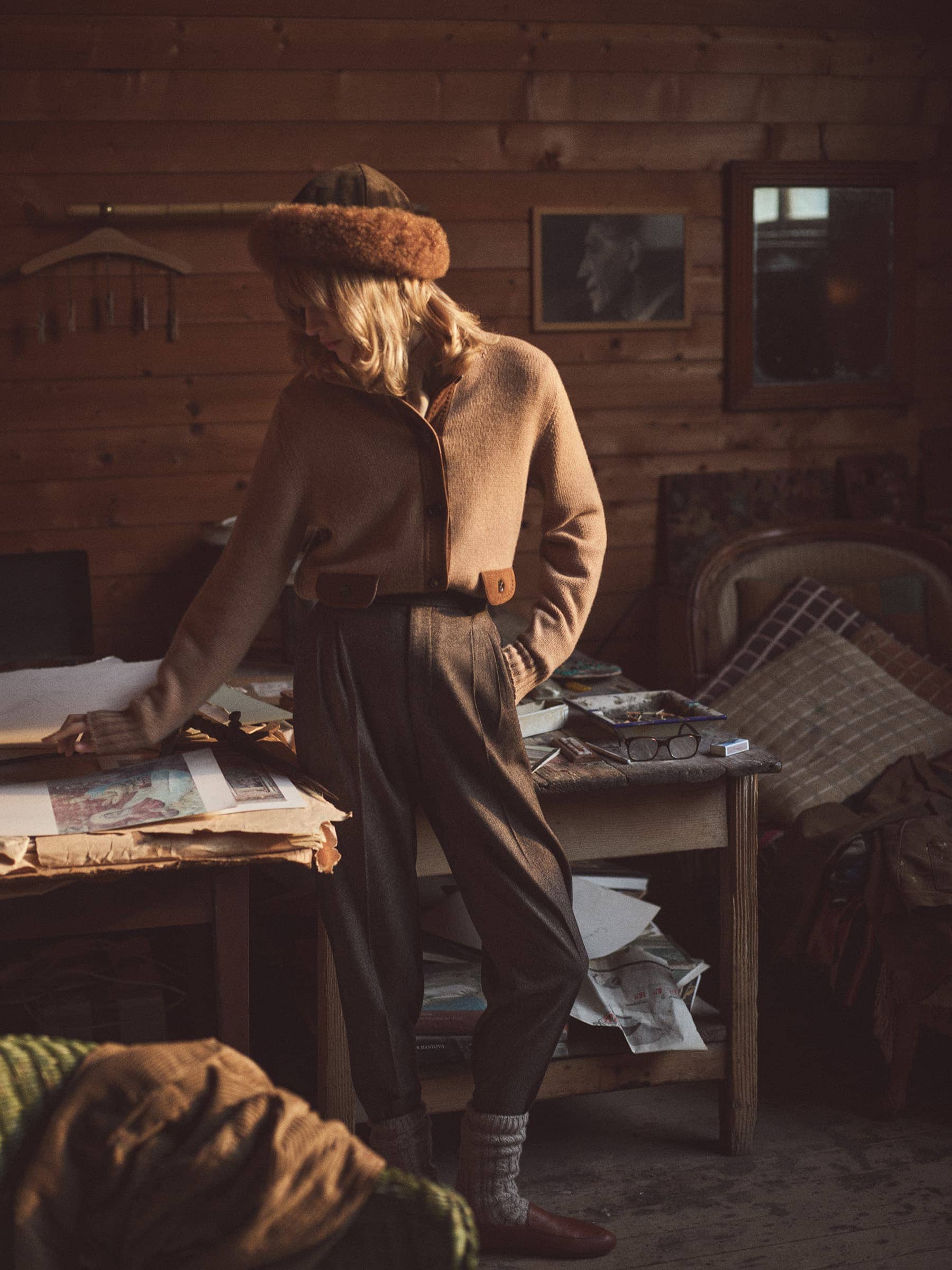 A blonde woman wearing a brown cardigan, wool hat and pants stands in a rustic wood paneled room with papers and objects on tables.