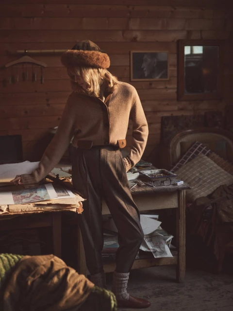 A blonde woman wearing a brown cardigan, wool hat and pants stands in a rustic wood paneled room with papers and objects on tables.