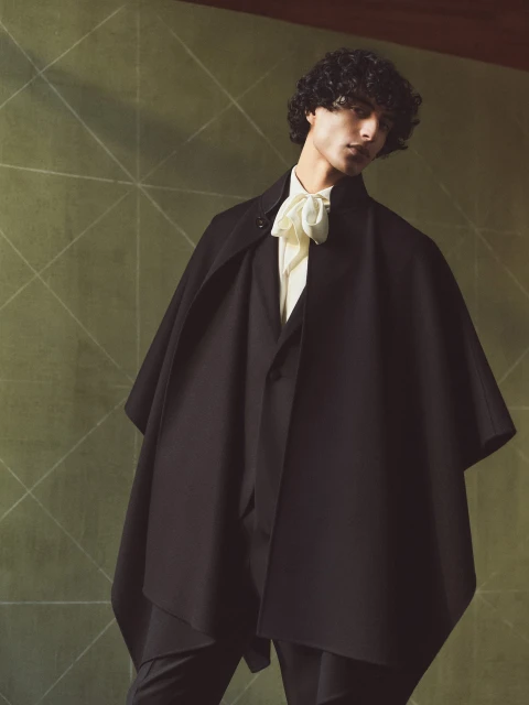 Studio shot of a handsome adult male model with curly hair posing in a black cape, vest, and pants, with a white ruffled cravat.