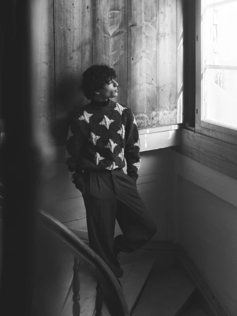 Young man stands on stairs by a window, wearing a sweater with stars and wide trousers in a black and white, indoor fashion shot.
