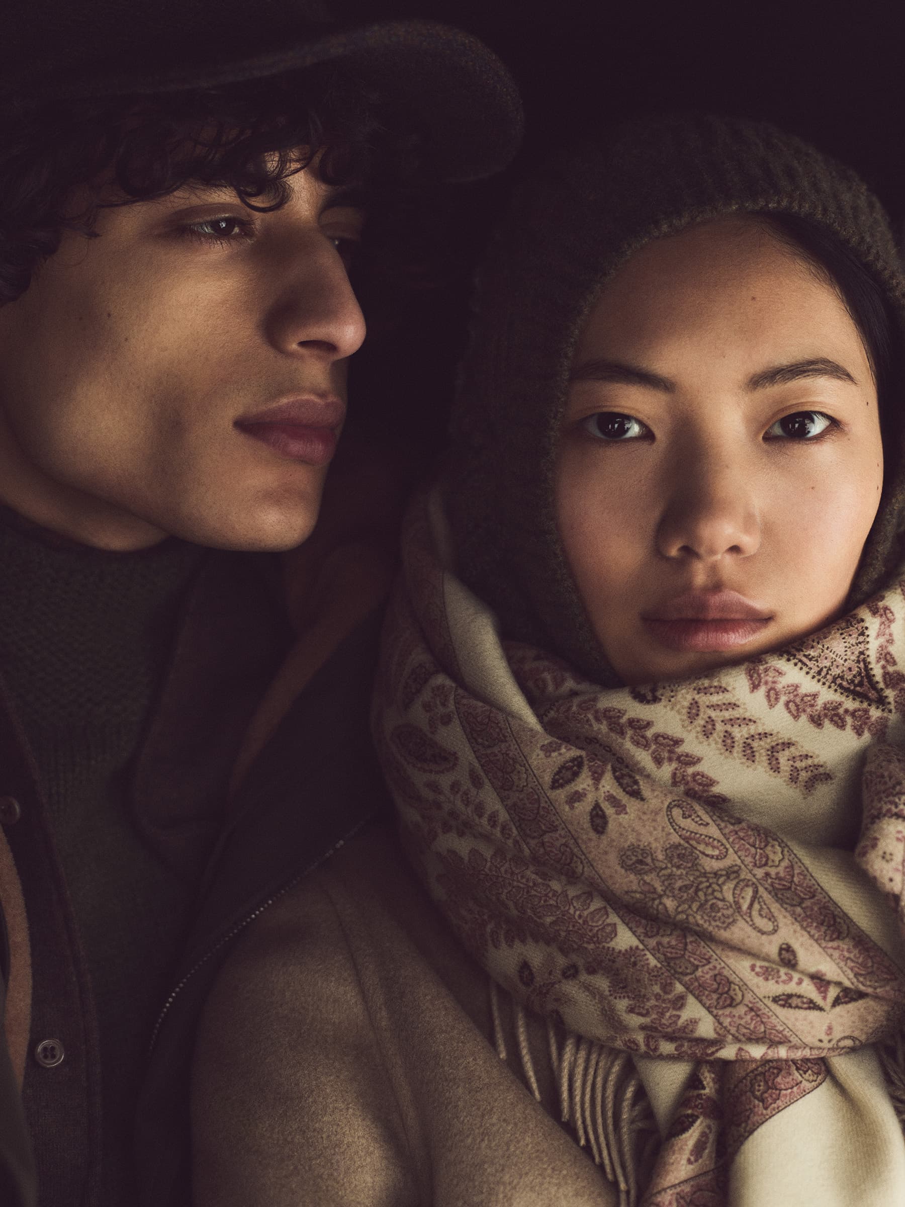 A couple poses intimately indoors in dark, ambient light. The woman is Asian with a patterned scarf, while the man wears a cap.