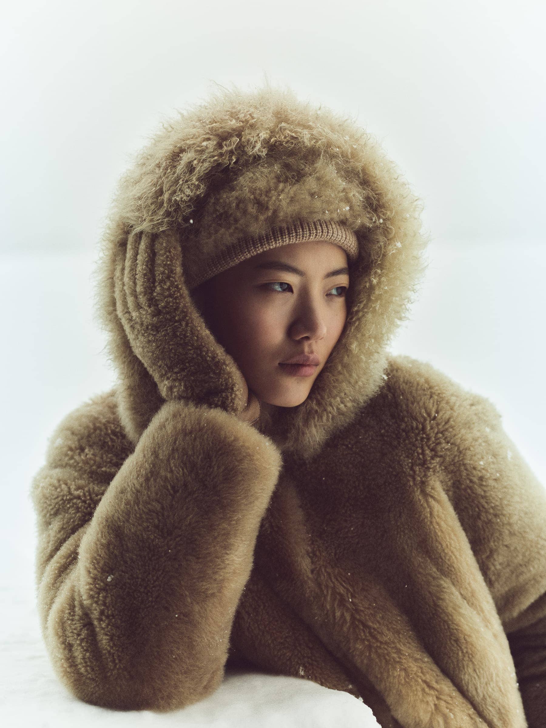 Portrait of a woman in a fur hat and coat looking to the side with her hand near her face in an outdoor, snowy environment.