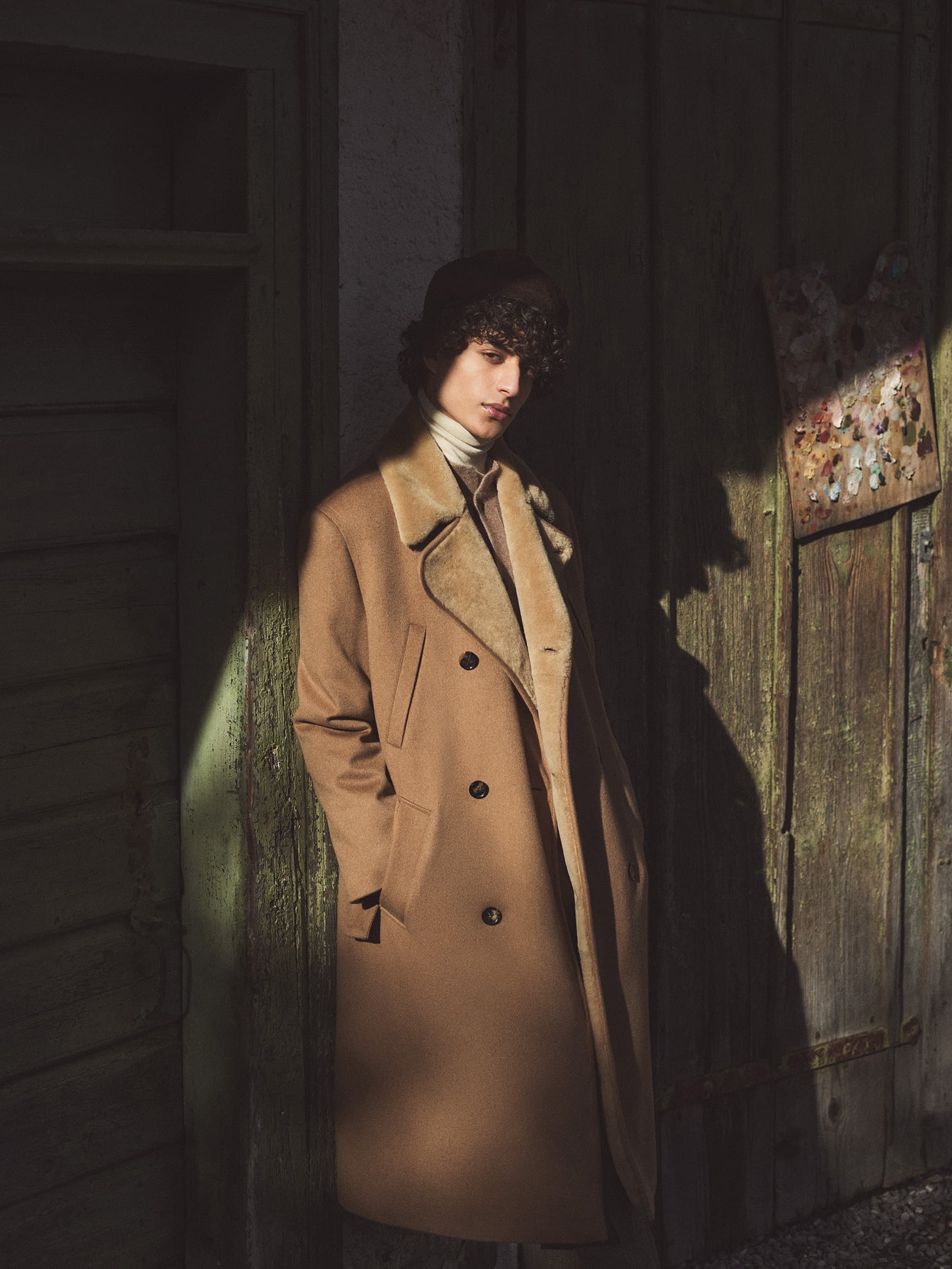 Adult male model in a tan overcoat and fur-trimmed hat poses in front of an aged green wooden door in outdoor lighting.