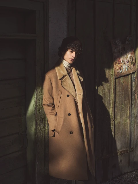 Adult male model in a tan overcoat and fur-trimmed hat poses in front of an aged green wooden door in outdoor lighting.