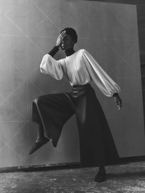 A Black woman in a cream-colored blouse and black pants strikes a pose with one hand covering her face, set against a textured wall.