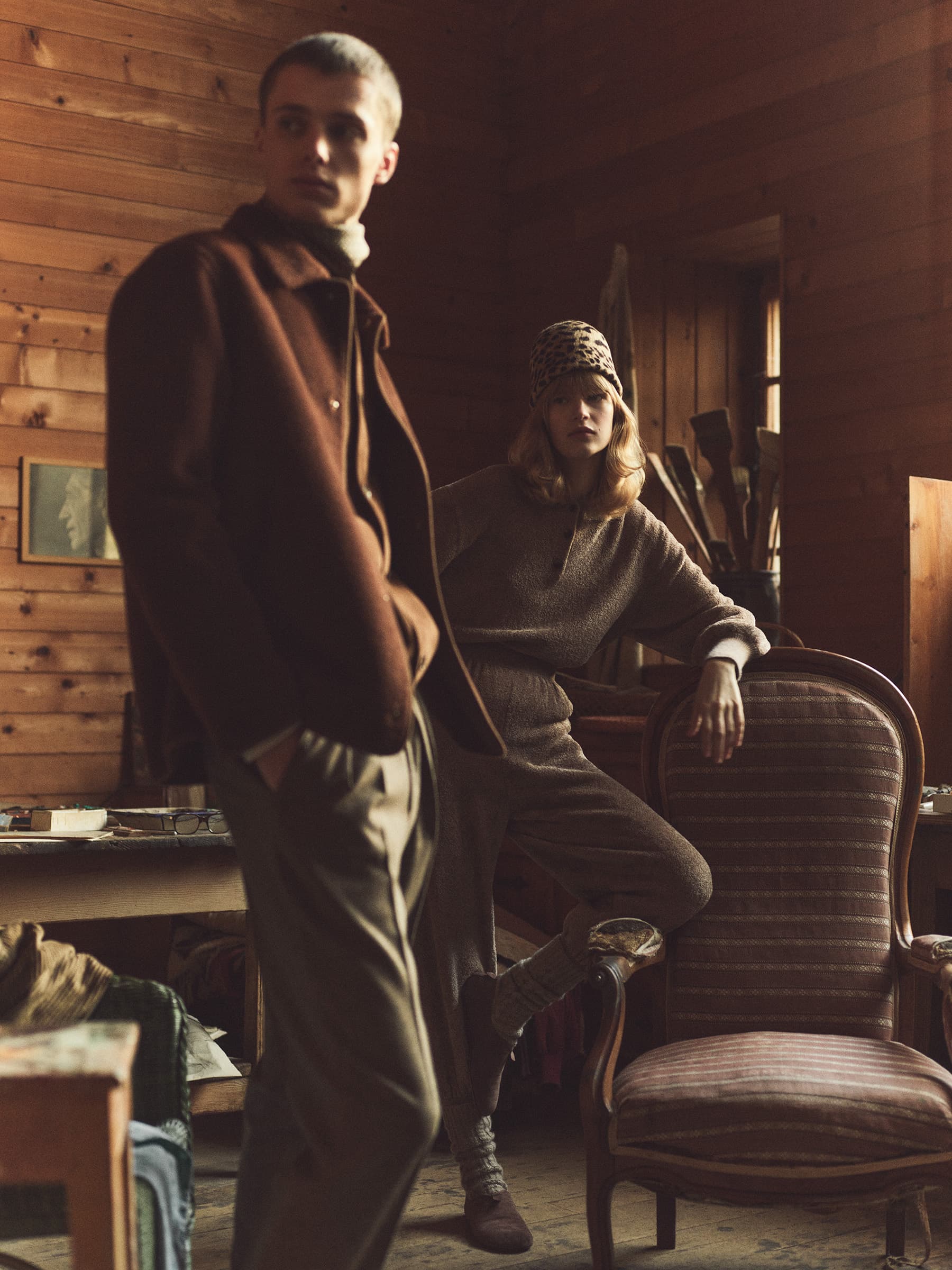Two fashion models pose in a rustic wooden interior wearing earth-tone knitwear and outerwear, with vintage furniture and soft lighting.