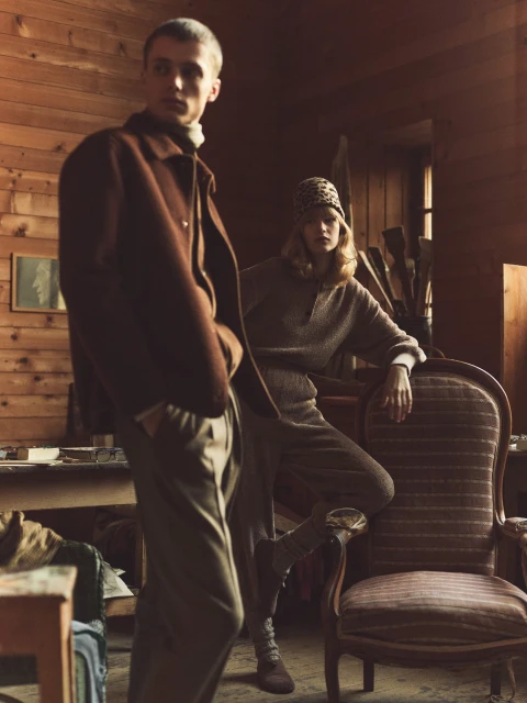 Two fashion models pose in a rustic wooden interior wearing earth-tone knitwear and outerwear, with vintage furniture and soft lighting.