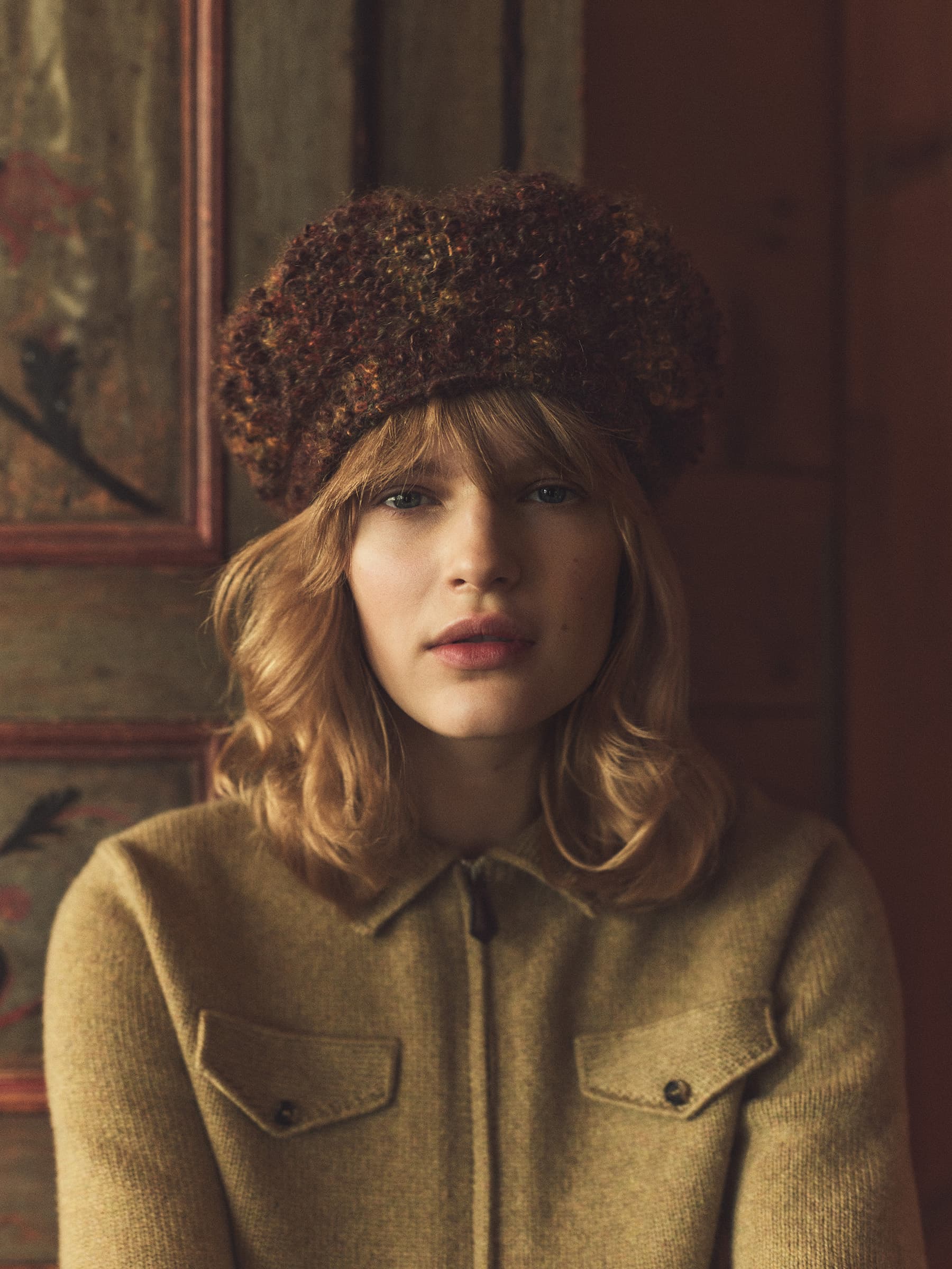 A young woman in a brown beret and a yellow jacket looks directly at the camera indoors.