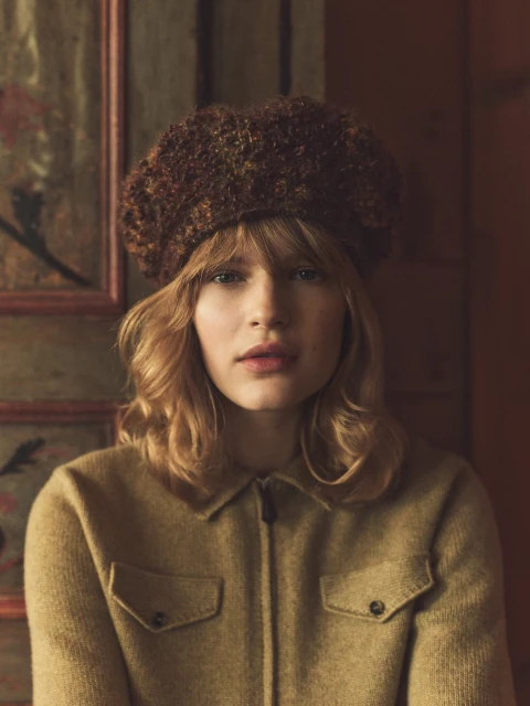 A young woman in a brown beret and a yellow jacket looks directly at the camera indoors.
