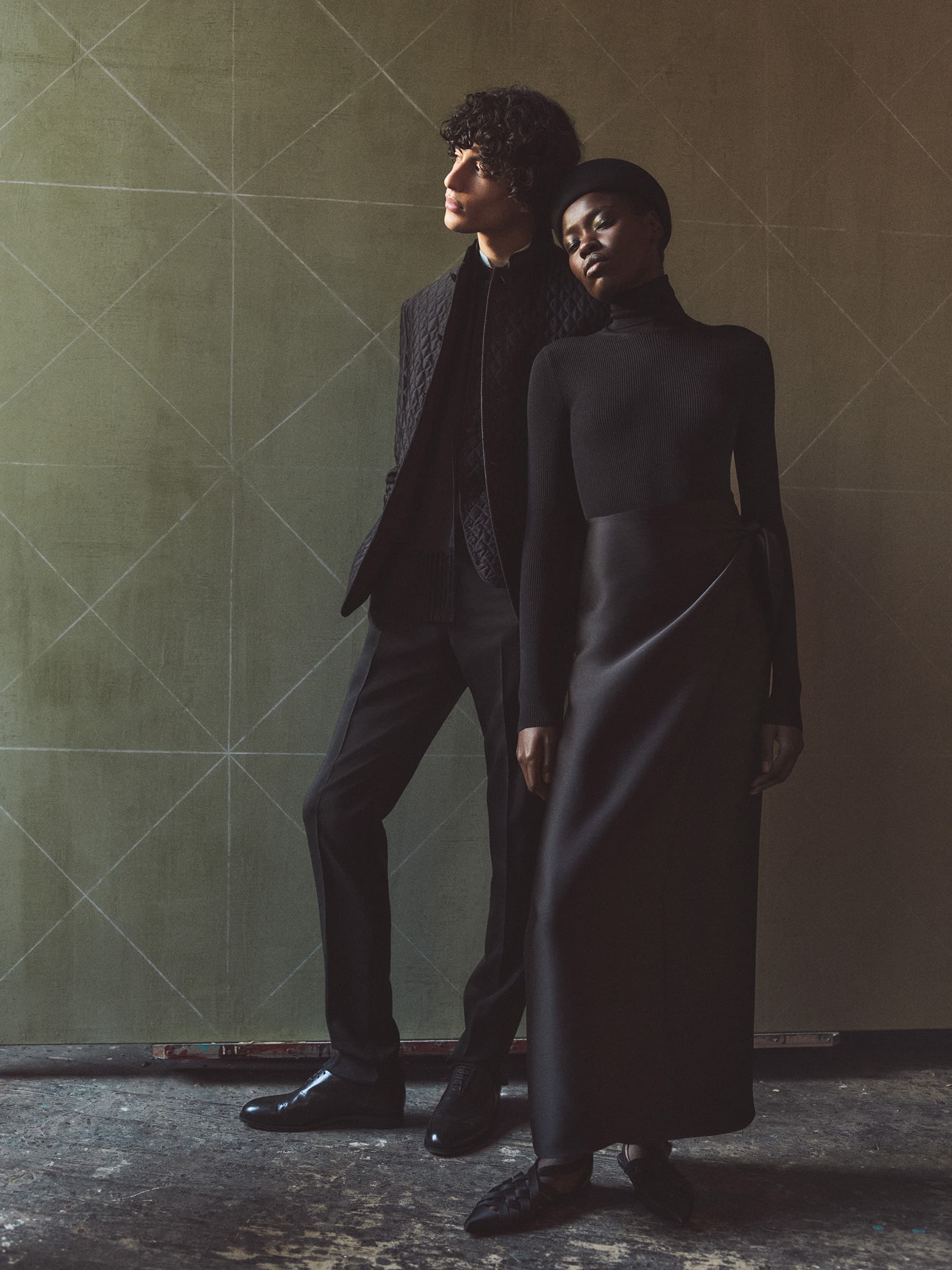 An adult male and an adult female pose together in a studio with geometric background, wearing dark formalwear and fashionable shoes.