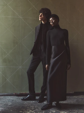 An adult male and an adult female pose together in a studio with geometric background, wearing dark formalwear and fashionable shoes.