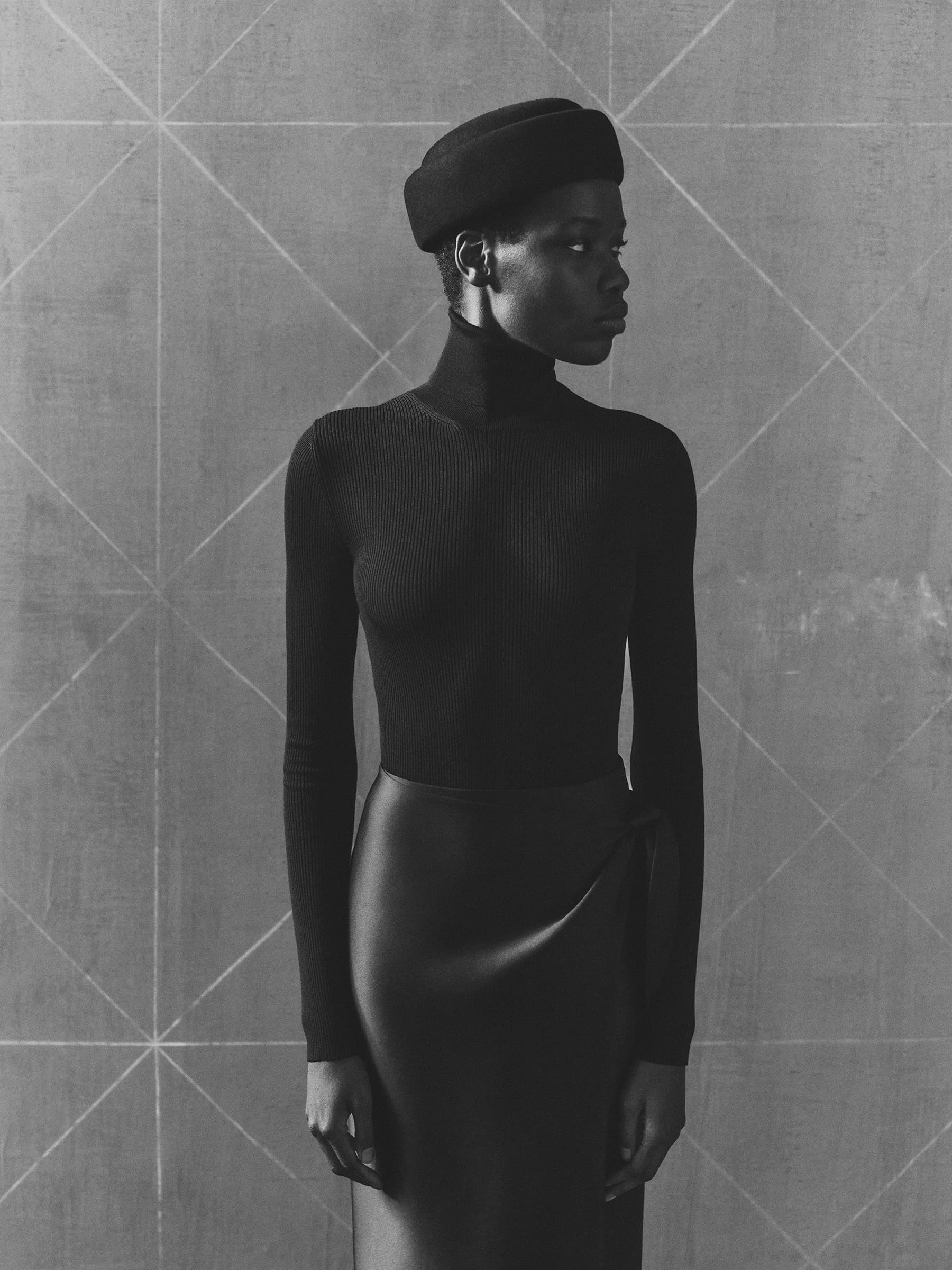 A Black woman stands in front of a gray wall with a geometric design wearing a black beret, turtleneck, and wrap skirt in a studio.