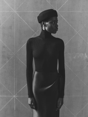 A Black woman stands in front of a gray wall with a geometric design wearing a black beret, turtleneck, and wrap skirt in a studio.