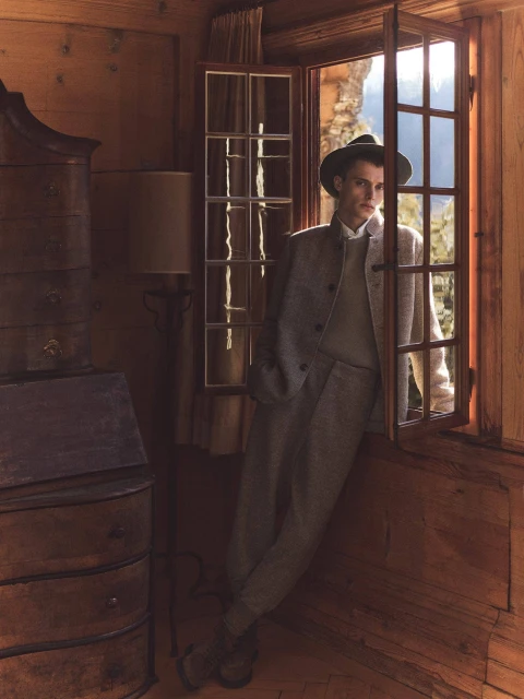 Indoor portrait of an adult male wearing a hat and tweed suit, leaning against an open window in a rustic wooden room.