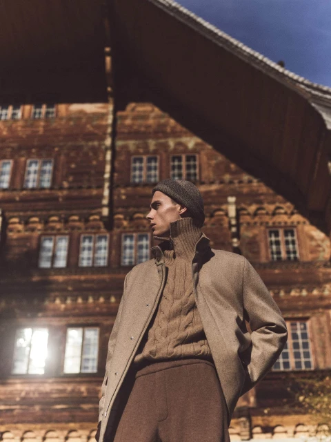 A young adult man in a beanie poses in outerwear, featuring a tan overcoat, cable-knit sweater, and brown trousers, against an alpine building backdrop.