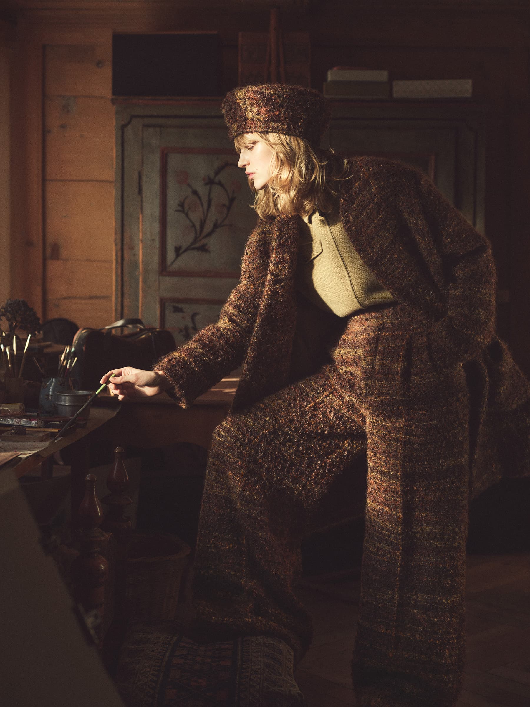 Indoor shot of a woman in a brown tweed suit and matching hat, leaning on a table with art supplies and neutral colored walls in the background.