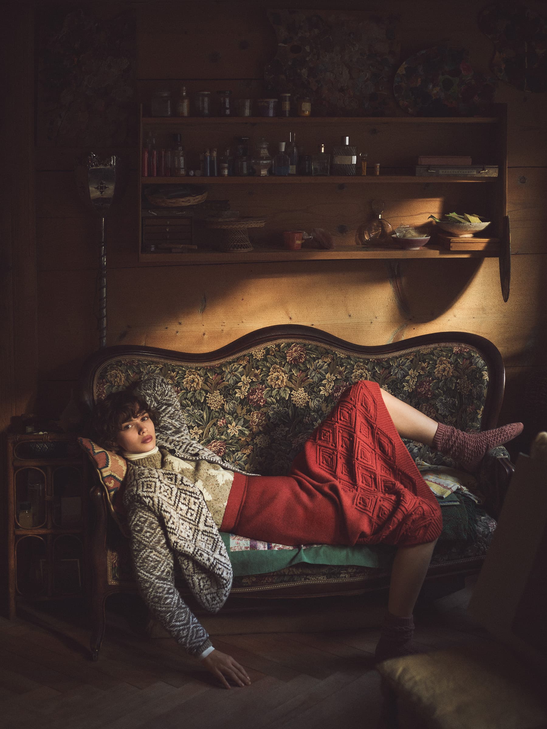 A woman lying on a patterned sofa wearing a patterned knit cardigan and red skirt in a dimly lit wood-paneled room