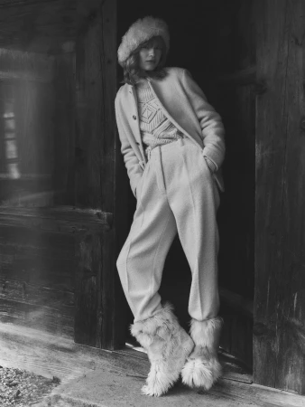 A woman wearing a light-colored winter ensemble including a textured wool jacket, sweater, matching trousers, a fuzzy hat, and fur boots.
