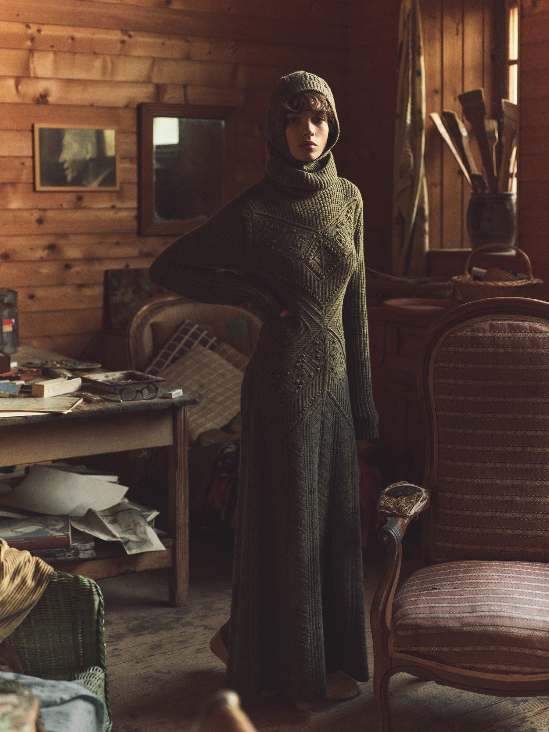 A woman in a long, olive green knit dress with a hood stands in a rustic room with wooden walls, antique furniture, and scattered papers.