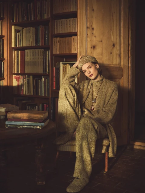 Indoor fashion portrait of a young adult female wearing a wool outfit and cap, seated in a chair, with a book case in the background.