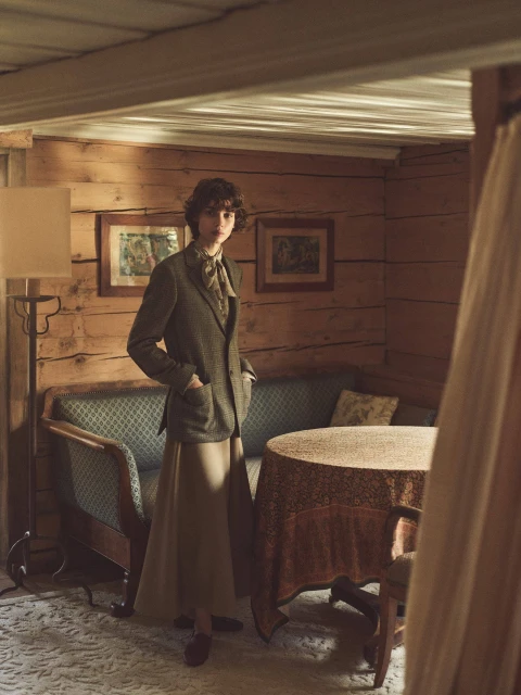 Woman in a blazer and long skirt stands inside a wood-paneled cabin, with antique furniture and soft, natural lighting. 