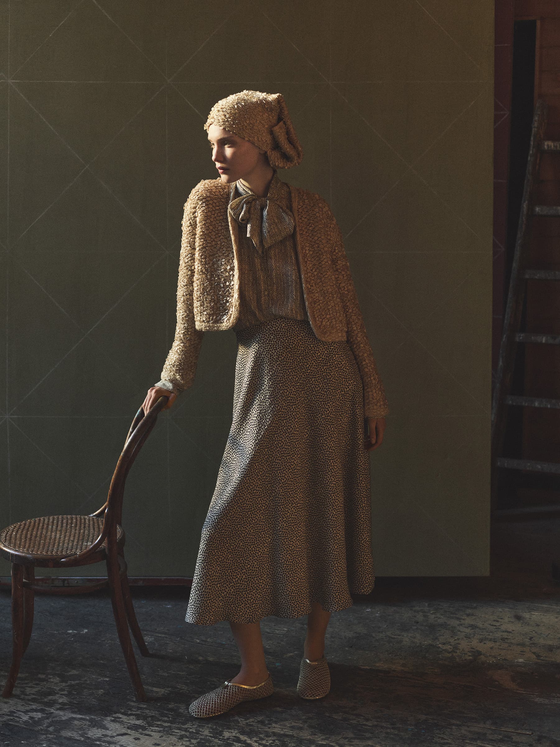 Model standing with hand on wooden chair wearing textured knit cardigan and patterned skirt