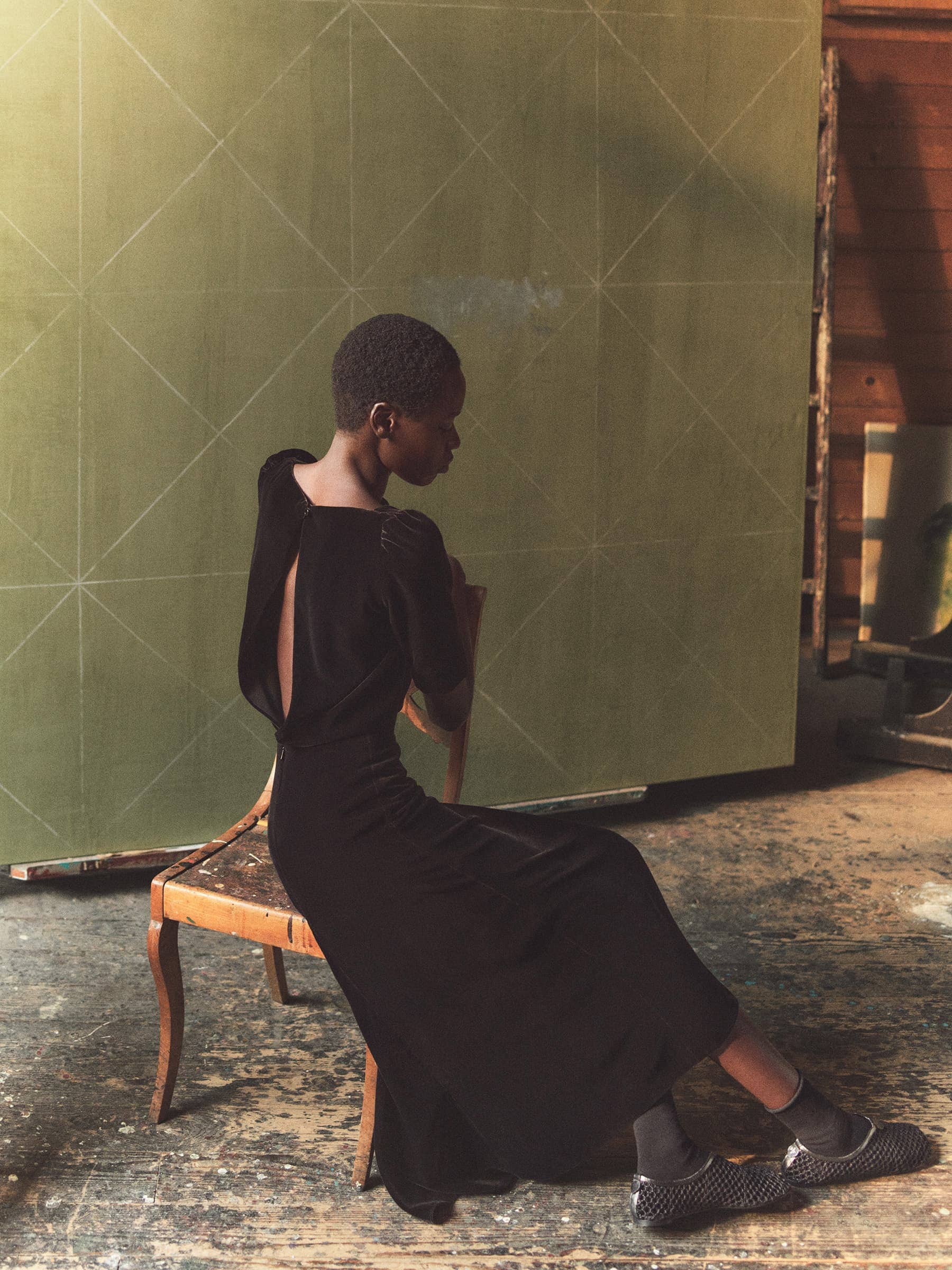 A dark-skinned woman with short hair poses sitting sideways on a chair. She is wearing a black dress with an open back in a studio setting.