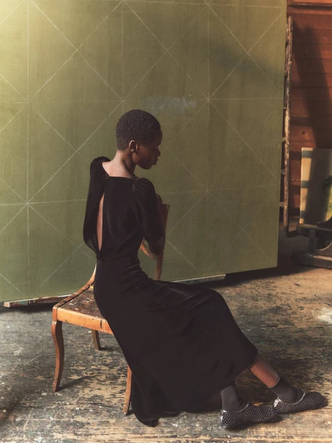 A dark-skinned woman with short hair poses sitting sideways on a chair. She is wearing a black dress with an open back in a studio setting.