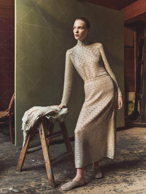 A slender woman in a patterned dress poses indoors with a wooden step stool, illuminated by diffused light, creating a fashion editorial image.