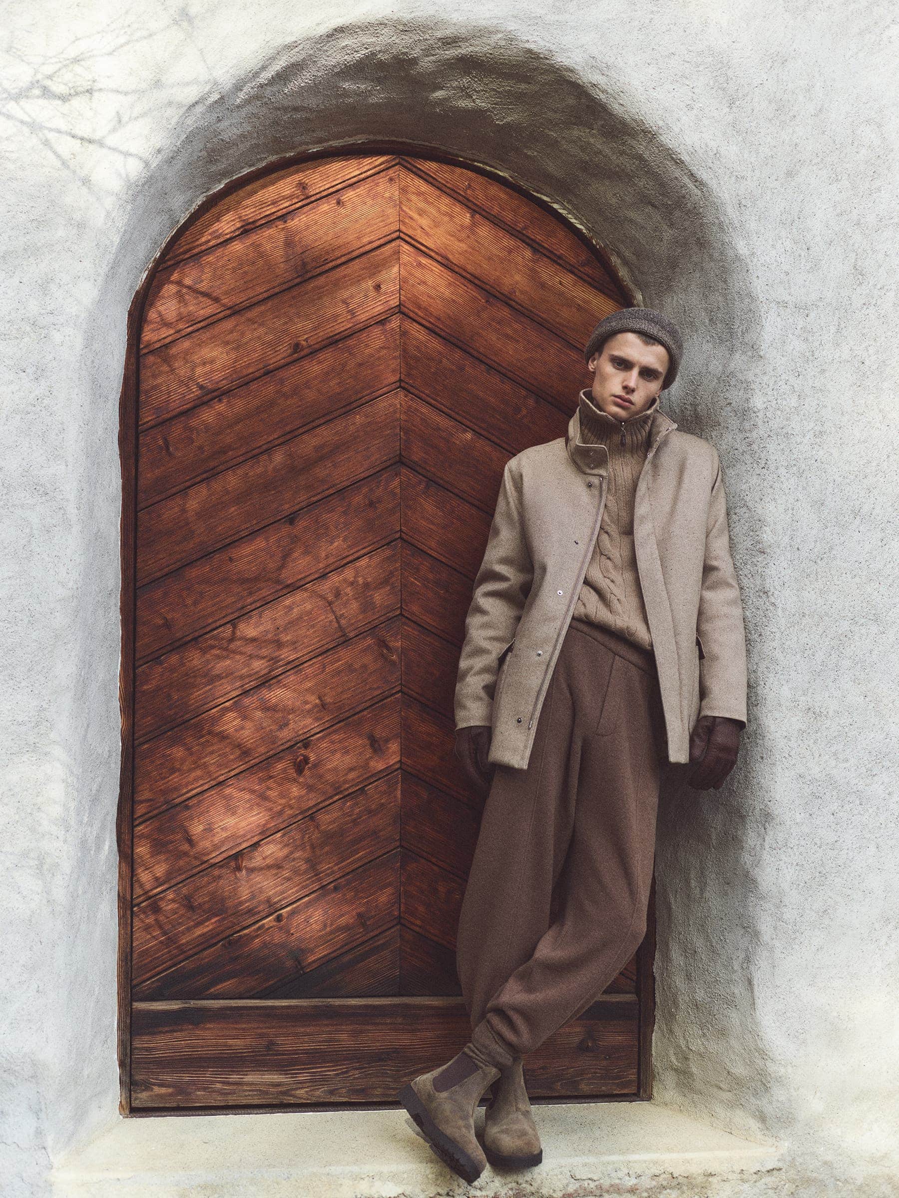 Man in a tan overcoat, cable-knit sweater, and beanie leaning against a rustic wooden door set in a stone archway. Outdoor fashion.