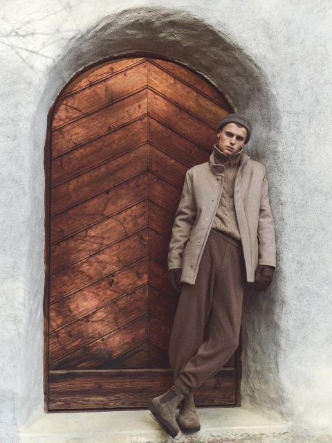 Man in a tan overcoat, cable-knit sweater, and beanie leaning against a rustic wooden door set in a stone archway. Outdoor fashion.