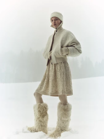 Model wearing a cream-colored textured zip jacket, matching skirt, fluffy winter boots, and knitted cap standing in the snow.