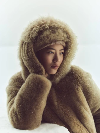 Model wearing a fluffy camel-colored shearling hooded coat and matching beanie against a plain light background