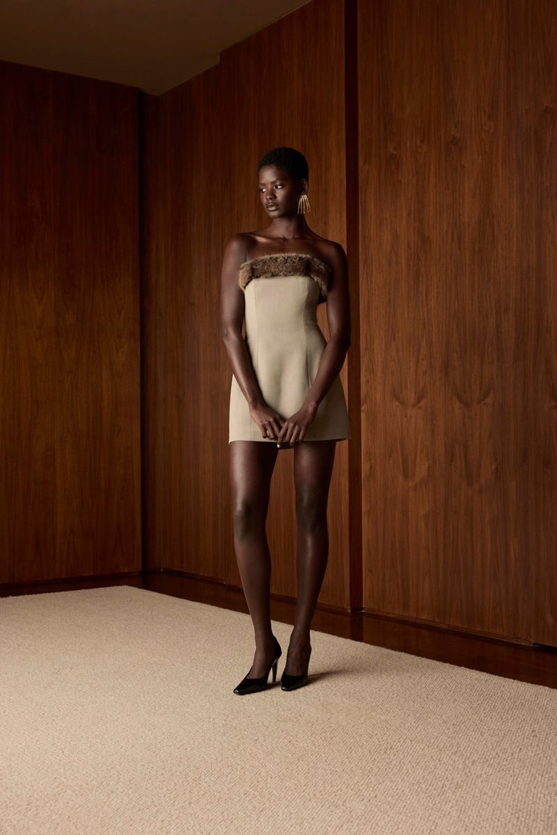 A black female fashion model poses in a strapless mini dress with brown trim, wearing black high heels in a minimalist studio.