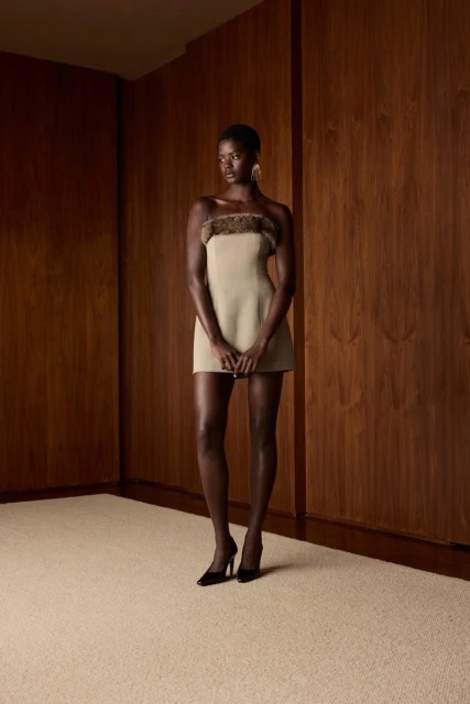 A black female fashion model poses in a strapless mini dress with brown trim, wearing black high heels in a minimalist studio.