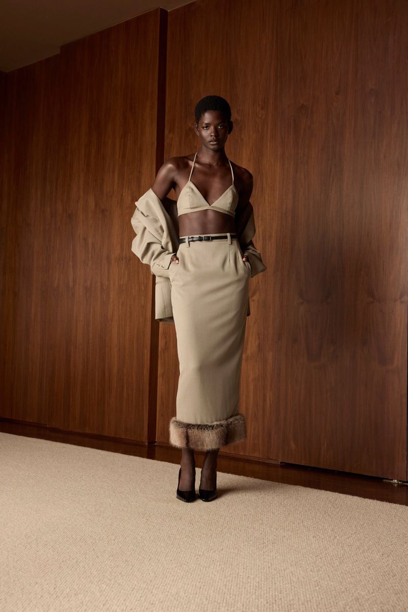Elegant Black woman poses indoors wearing beige bra top, skirt with fur trim, and blazer with wooden wall and carpet backdrop.