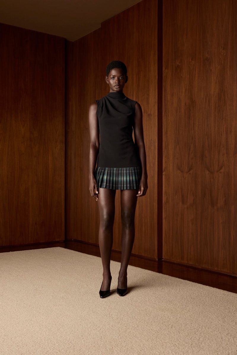 Elegant woman with dark skin in black top and plaid miniskirt standing against brown wood-panelled wall, minimalist style in studio.