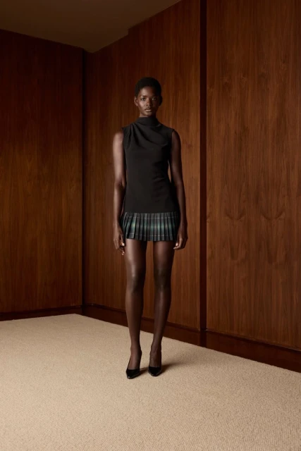 Elegant woman with dark skin in black top and plaid miniskirt standing against brown wood-panelled wall, minimalist style in studio.