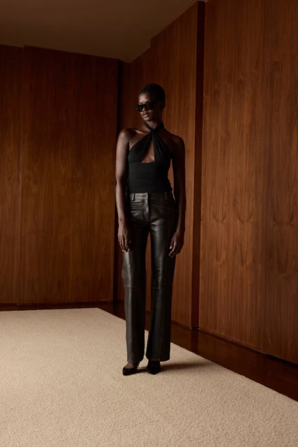 A woman with sunglasses stands in front of a wood paneled wall, wearing a black top and leather pants in an indoor setting.